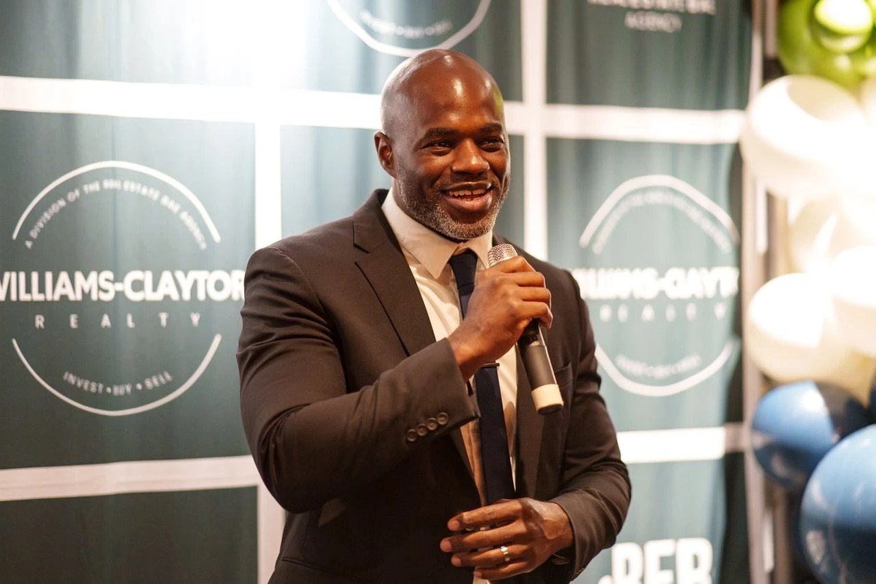 John Lewis smiling with a microphone in front of a Williams-Claytor Realty backdrop